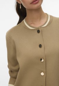 Beige cardigan met ribbeltextuur, voorzien van een contrasterende witte gestreepte kraag, met grote gouden knopen aan de voorkant.