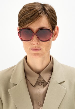 Persona con capelli castano chiaro corti indossa grandi occhiali da sole esagonali di colore rosso scuro, camicia beige con colletto e blazer beige coordinato.