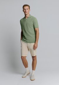 Groen polohemd met textuur en korte mouwen, gecombineerd met beige korte broek en witte sneakers, tegen een neutrale achtergrond.