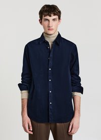 Camicia blu navy con bottoni in un tessuto morbido e testurizzato. Presenta un colletto classico e polsini arrotolati, indossata sopra un dolcevita beige.