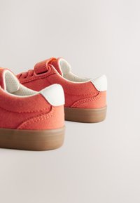 Pomarańczowe zamszowe trampki z białymi akcentami, zaokrąglonym noskiem, wiązane na sznurówki, z fakturowaną tkaniną oraz beżową gumową podeszwą. Posiadają pasek na rzep.