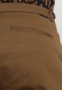 Pantalons marron avec une texture lisse, dotés d'une poche arrière unique avec une ouverture horizontale et des détails de couture subtils.