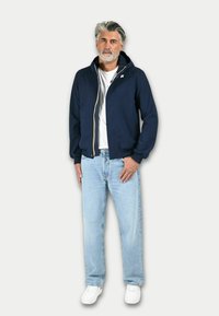 Felpa con zip color navy, con tasche laterali e polsini a coste, indossata sopra una t-shirt bianca e jeans a gamba dritta di colore blu chiaro. Sneakers bianche.