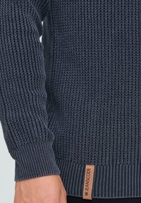 Tume sinine kudunud särk, mille varrukad ja äär on ribitud ning millel on väike beež silt, millel on kirjas "JEANSCODE", vöökohal mustade pükste peal.