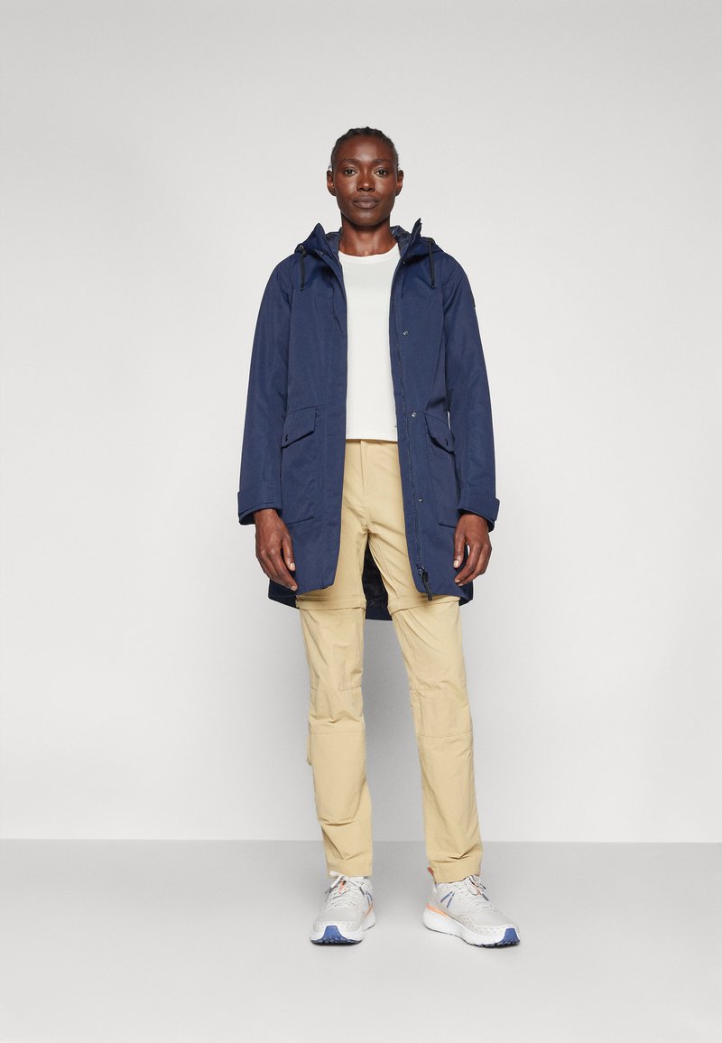 Parka étanche bleu marine avec capuche, fermeture éclair frontale et deux grandes poches, associé à un pantalon cargo beige et des baskets grises.