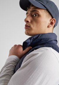 Marineblau gestepptes Westchen mit hohem Kragen, getragen über einem weißen Langarmshirt, mit einem Reißverschluss, vor einem neutralen Hintergrund.
