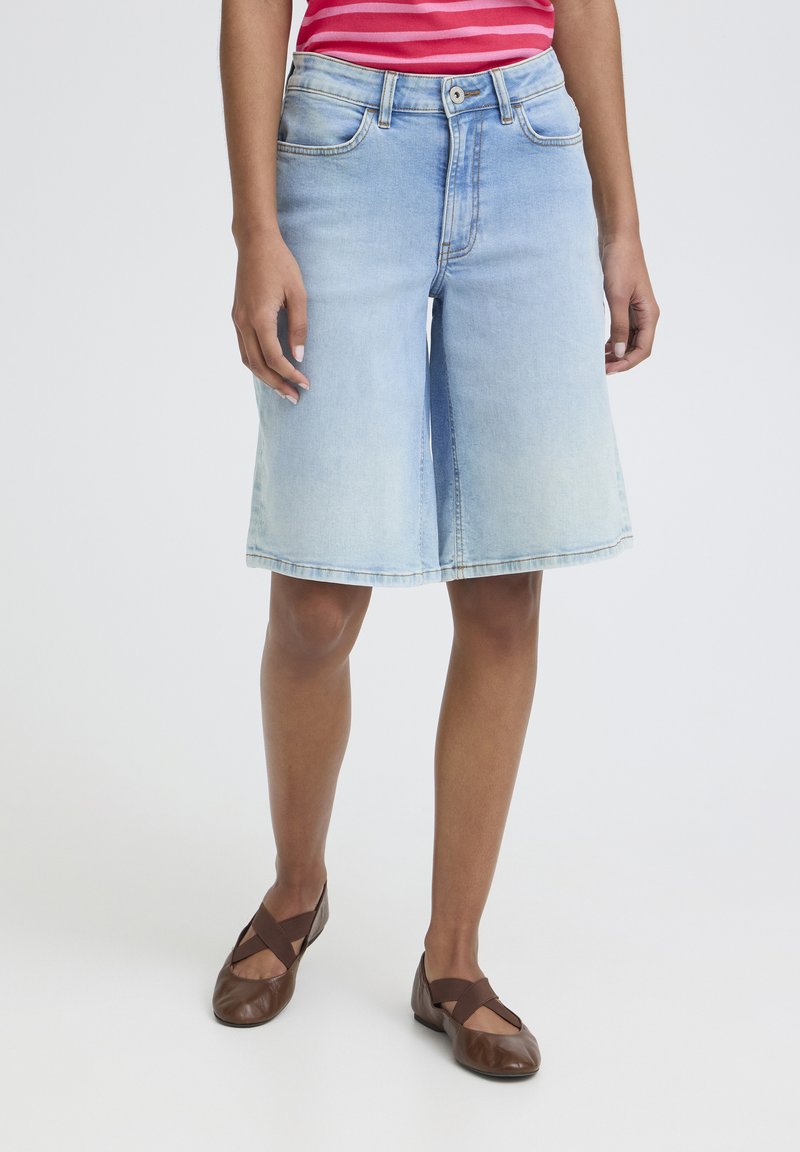 Personne portant une jupe en denim bleu clair jusqu'aux genoux, un haut rayé rose, et des chaussures plates marron à brides croisées, debout sur un fond blanc.