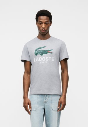 Homme portant un t-shirt Lacoste gris clair avec le logo crocodile vert et le texte "LACOSTE PARIS", assorti à un jean bleu clair.