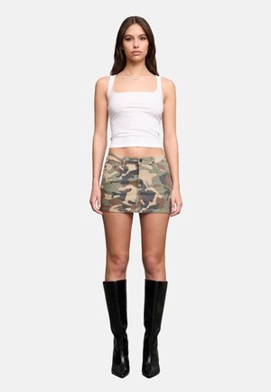 Donna con canotta bianca, minigonna mimetica e stivali neri al ginocchio, in piedi davanti a uno sfondo chiaro e uniforme.