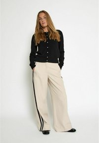 Black cardigan with button front, cream wide-leg trousers featuring black side stripes, and black shoes on a plain background.