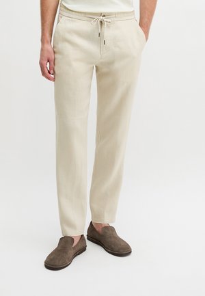 Man die beige broek met trekkoord en bruine suède instapschoenen draagt, staand met handen in de zakken tegen een witte achtergrond.