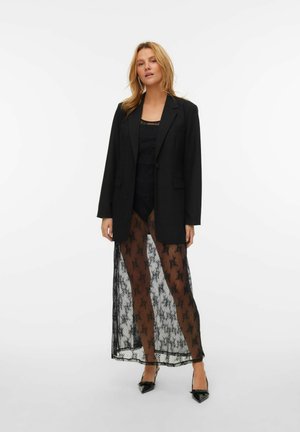 Zwarte blazer, doorschijnende zwarte kanten jurk met bloemenpatroon, over een zwarte top, en zwarte puntige hakken. Strakke lijnen en een elegante uitstraling.