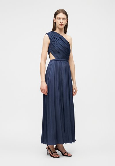 Femme portant une robe de soirée plissée bleu marine à une épaule, avec une découpe latérale, et des sandales à talons noires à lanières, debout devant un fond blanc.
