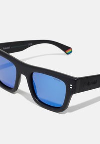 Occhiali da sole neri a forma quadrata con lenti blu. Montatura nera testurizzata con il logo Polaroid. Accentatura arcobaleno sulla tempia.