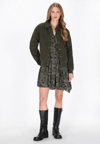 Zielona dżinsowa kurtka na tle ciemnej sukienki w kwiaty. Sukienka ma warstwowy design. Modelka nosi wysokie czarne skórzane botki z klamrami.