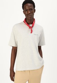Tricou gri cu mânecă scurtă dintr-un amestec de bumbac, având un mic logo pe piept. Asortat cu pantaloni bej și un batic roșu în jurul gâtului.