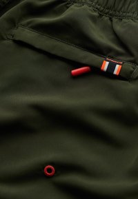 Groene stoffen shorts met een gladde textuur, voorzien van een rode rubberen label en een meerkleurig merketiket op de tailleband, met een drukknoopdetail.