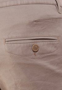 Pantalons en tissu beige avec une poche arrière dotée d'un design rectangulaire cousu et d'un bouton rond de couleur claire. Texture lisse, coupe ajustée.