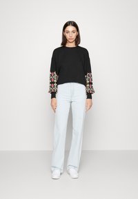 Sweatshirt preto curto com padrões bordados coloridos nas mangas. Usado com jeans flare azuis claros e ténis brancos.