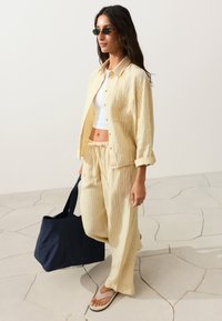 Femme portant une chemise rayée jaune et un pantalon avec un top court blanc et des tongs, portant un grand sac cabas bleu marine, marchant sur un sol en pierre.