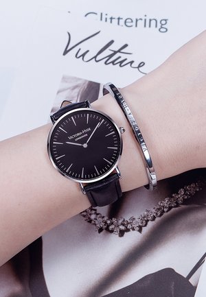 Reloj con correa de cuero negro, caja redonda de acero inoxidable, esfera negra, marcadores de hora elegantes, acompañado de una pulsera de bangle de plata con un logo grabado.
