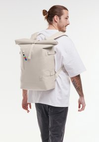 Sac à dos beige à rabat, fabriqué en matériau durable, avec des bretelles réglables, une poche zippée à l'avant et un logo imprimé sur le côté.