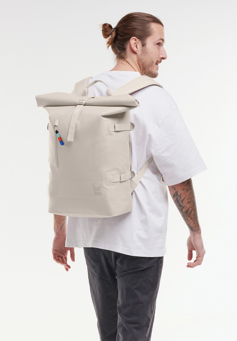 Sac à dos beige à rabat, fabriqué en matériau durable, avec des bretelles réglables, une poche zippée à l'avant et un logo imprimé sur le côté.