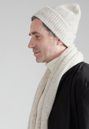 Berretto lavorato a maglia di un beige chiaro con texture a coste, abbinato a una sciarpa a pois coordinata e a una giacca nera. Sotto una maglia a collo alto.