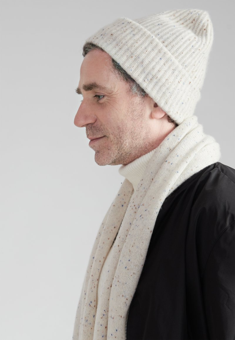 Light beige knit beanie with ribbed texture, paired with a matching speckled scarf and a black jacket. Turtleneck underneath.