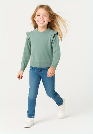 Vaaleanvihreä neulottu villapaita, jossa on röyhelöillä varustetut olkapäät, yhdistettynä sinisiin farkkuihin ja valkoisiin lenkkareihin. Lapsi kävelee hiukset lainehtien.