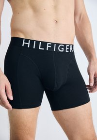 Mannlig overkropp iført svarte boxer briefs med "HILFIGER" trykt i hvitt på linningen mot en ensfarget bakgrunn.