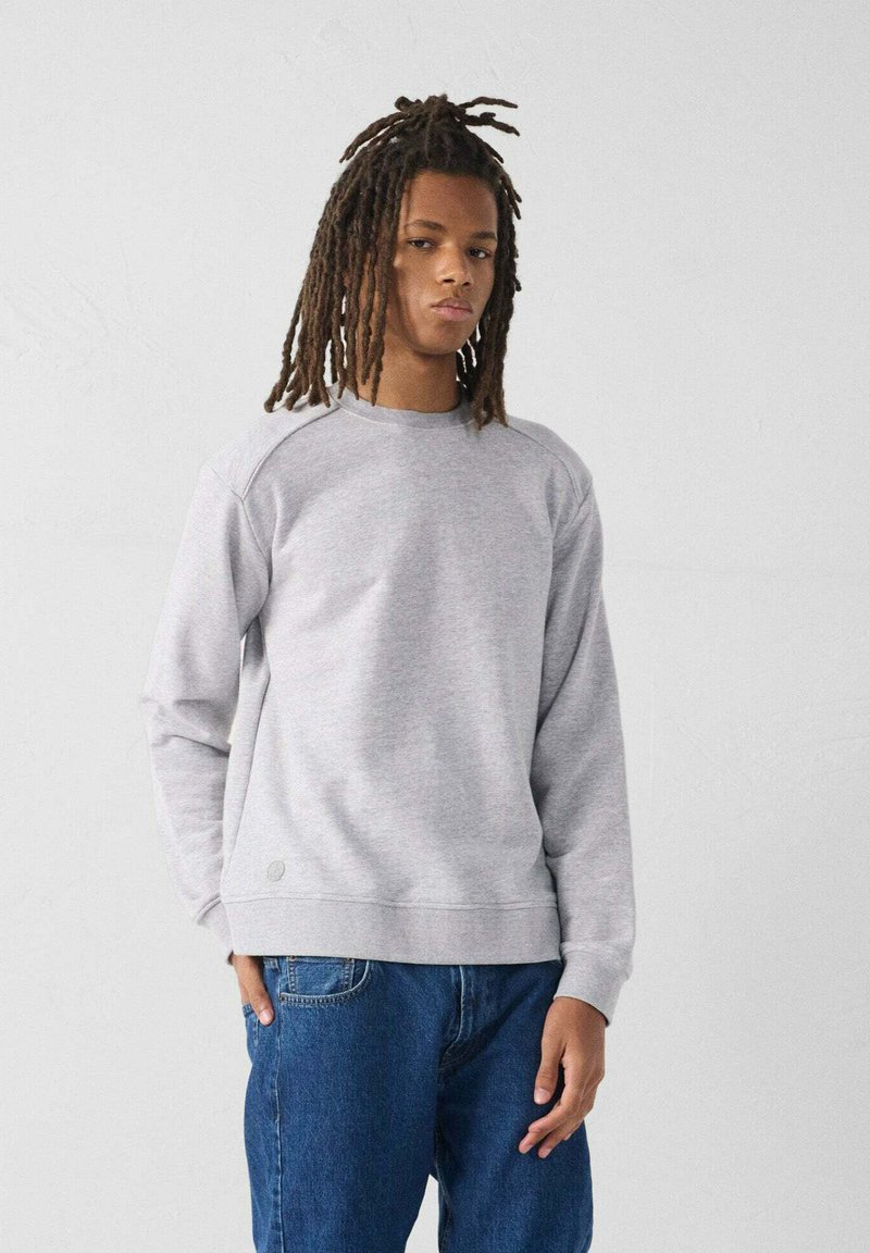 Felpa grigia con collo rotondo, maniche raglan e polsini a costine. Abbinata a jeans blu. Branding minimale sulla parte inferiore anteriore.