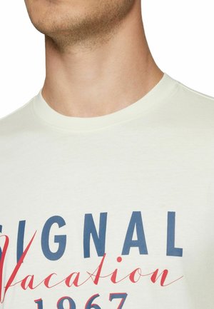 Nærbillede af en persons nakke og øverste del af brystet med en hvid t-shirt med teksten "SIGNAL vacation 1967" i blå og rød.