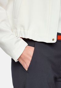Weiße, kurz geschnittene Jacke mit gerafftem Saum, kombiniert mit dunkler Hose. Die Jacke hat eine glatte Textur; die Hose ist schlank mit seitlichen Taschen.