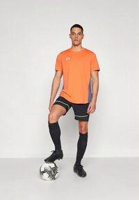 Camiseta deportiva naranja con paneles laterales grises, pantalones cortos negros con ribete neón y tacos de fútbol negro, de pie sobre un balón de fútbol blanco y negro.