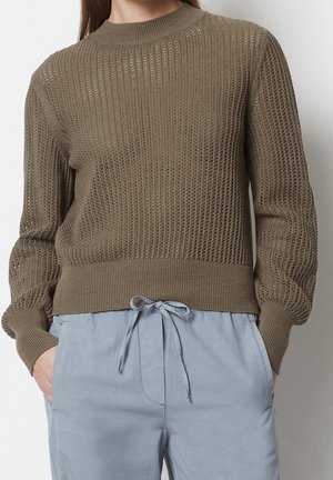 Femme portant un pull en maille marron texturé associé à un pantalon bleu clair à cordon, mains dans les poches, sur un fond uni.
