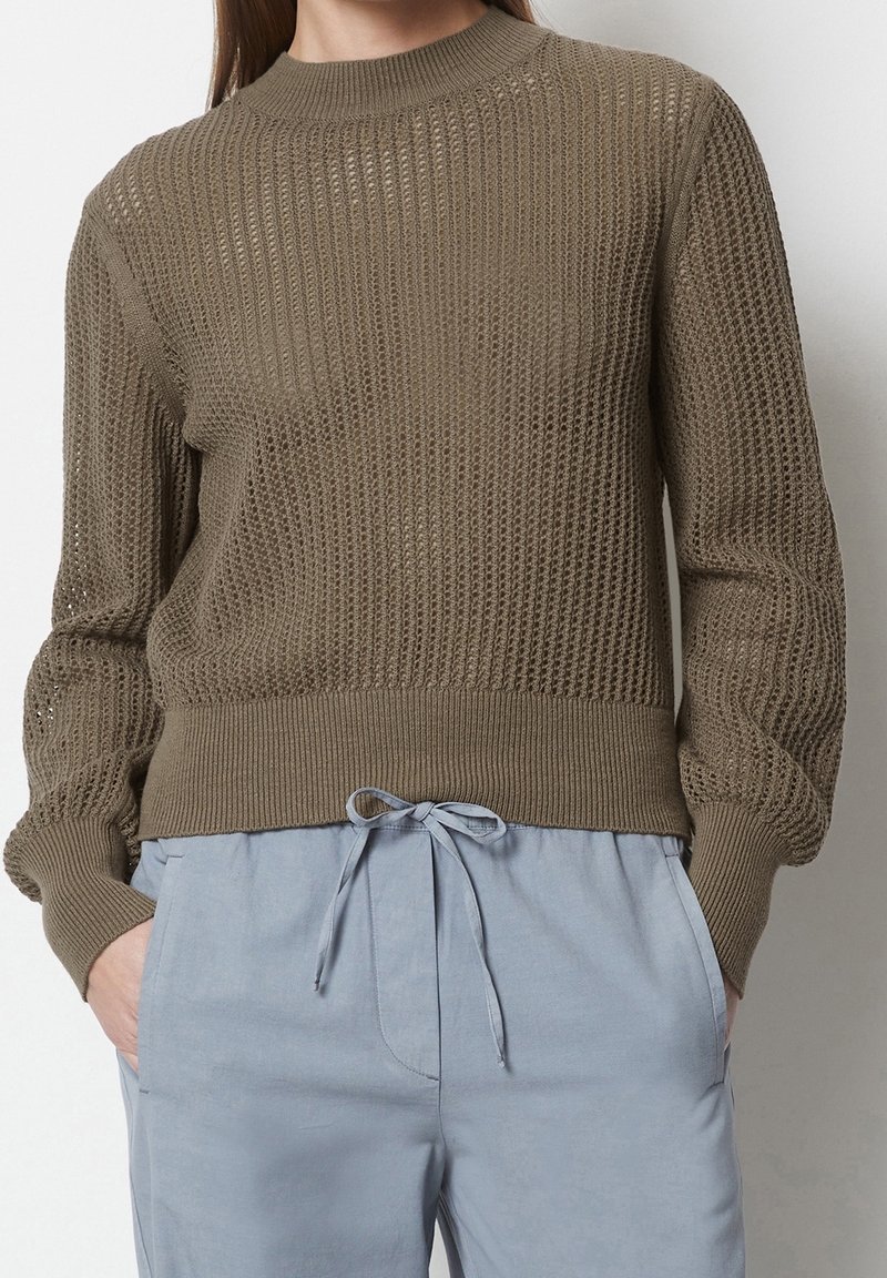 Femme portant un pull en maille marron texturé associé à un pantalon bleu clair à cordon, mains dans les poches, sur un fond uni.