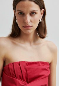 Czerwona sukienka bez ramiączek z plisowanymi akcentami, wykonana z gładkiego materiału. Modelka pokazuje delikatny makijaż i srebrne okrągłe kolczyki na jednolitym tle.