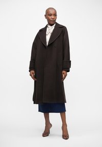 Σκούρο καφέ μάλλινο παλτό με φαρδιά πέτα, μεγάλα κουμπιά και τσέπες με παύλες. Λείος υφή, μήκους μέχρι το γόνατο, φορεμένο με τζιν φούστα.