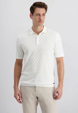 Homme portant un polo à manches courtes blanc avec un motif en losange et un pantalon beige clair, debout devant un fond uni.