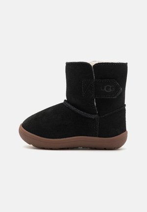 Bottine en suede noir avec un bout arrondi, semelle en caoutchouc beige et doublure en polaire. Présente une étiquette sur le côté et un détail de cordon décoratif.