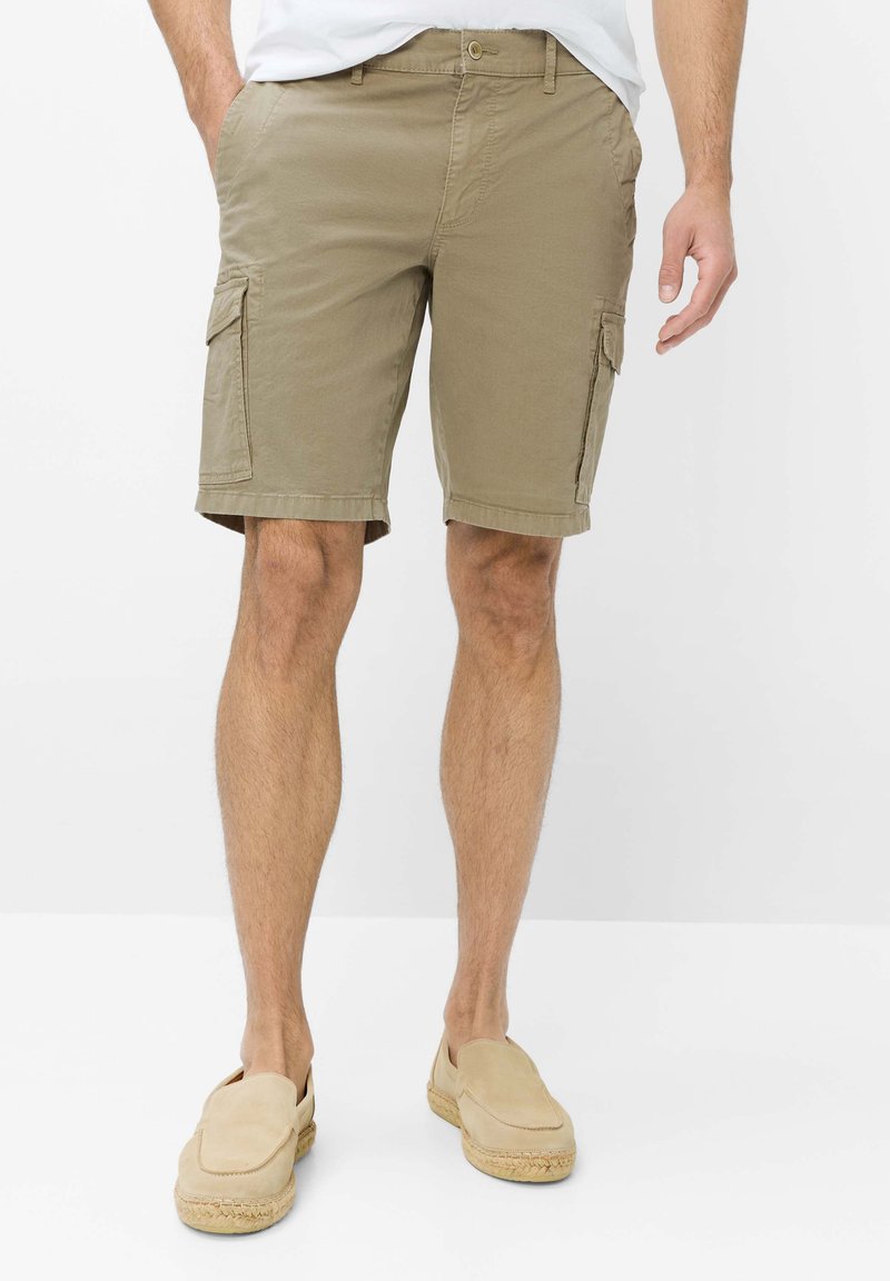 Homme portant un short cargo beige et des chaussures beige sans lacets, debout les mains dans les poches devant un fond uni.