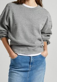 Sudadera gris con un diseño recortado y hombros caídos, con adornos negros en las mangas. Combinada con una falda de mezclilla clara.