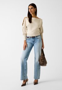 Beżowy dziergany sweter z akcentami na ramionach, w zestawie z jasnoniebieskimi jeansami o prostych nogawkach, brązową torebką i brązowymi butami na wysokim obcasie.