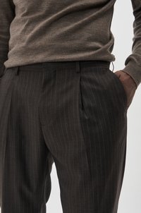 Dunkelgraue, gestreifte Hose mit taillierter Passform, Falten an der Vorderseite und seitlichen Taschen, aus glattem Stoff, kombiniert mit einem braunen Pullover.