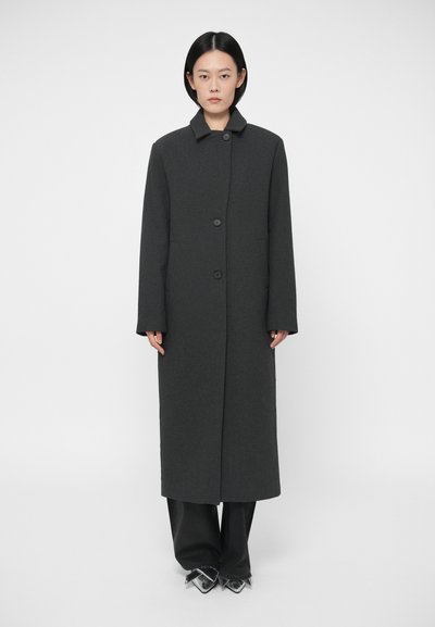Femme portant un long manteau gris foncé avec boutons, un pantalon noir large et des chaussures argentées pointues, se tenant devant un fond blanc uni.