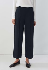 Kvinna som bär svarta vida capribyxor, vit långärmad topp och svarta spetsiga platta skor, stående på ett vitt golv.