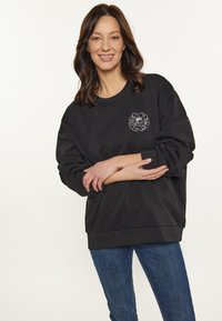 Sudadera negra con un corte relajado, puños acanalados y un diseño floral bordado en el lado izquierdo del pecho. Combinada con vaqueros azules.