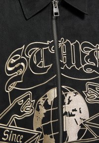 Musta vetoketjullinen takki, jossa on teksturoitu materiaali, kullanvärinen kirjonta, jossa maapallo, sekä graafisia elementtejä, kuten tyyliteltyjä kirjaimia ja käsiä.