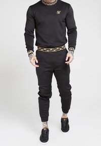 Svart träningsset med lång ärm och smalnande byxor, med ett guld geometriskt mönster på kragen och midjebandet. Svarta sneakers ingår.